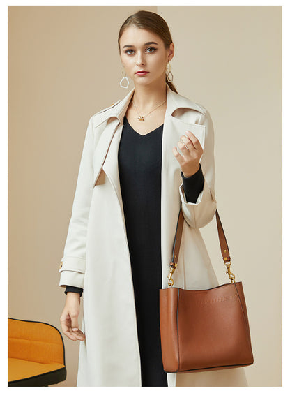 Model in cream coat and black dress carrying tan bucket bag at her side