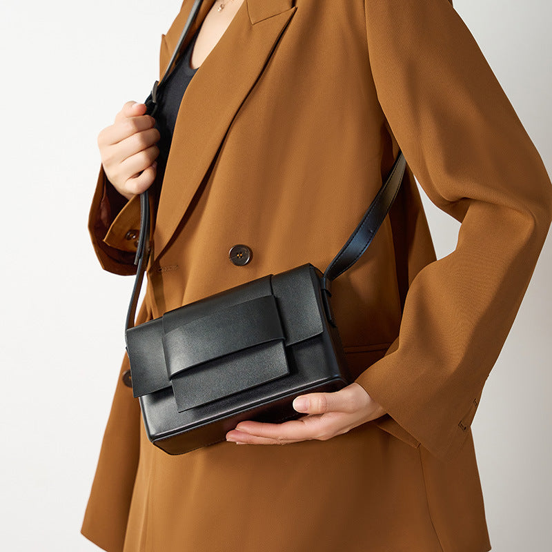 Black leather bow flap crossbody bag held in hand against a tan blazer
