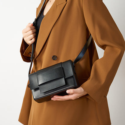 Black leather bow flap crossbody bag held in hand against a tan blazer