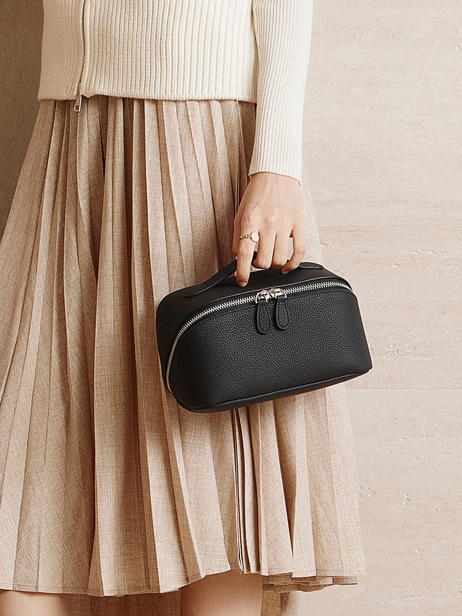 Black pebbled leather cosmetic pouch carried by top handle against a pleated beige skirt