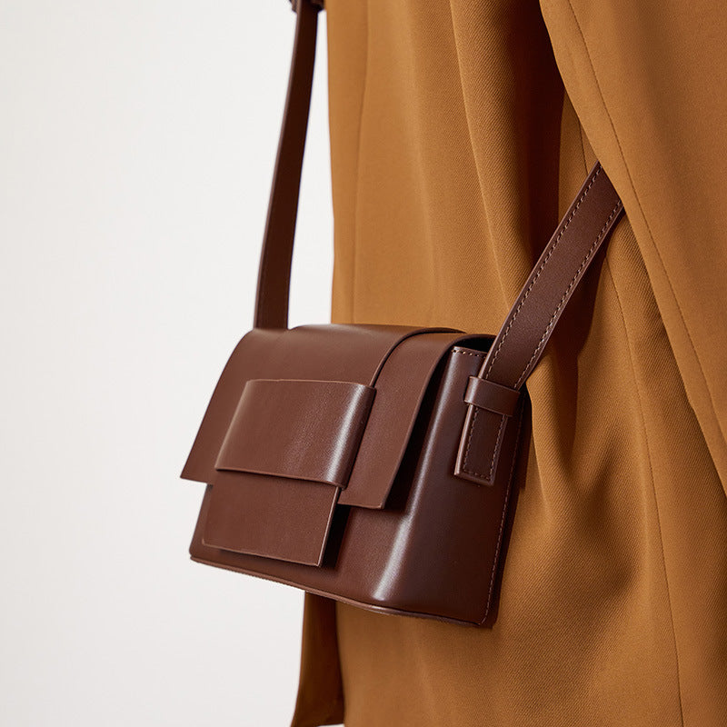 Coffee brown leather crossbody bag with bow detail worn at the hip with a tan coat