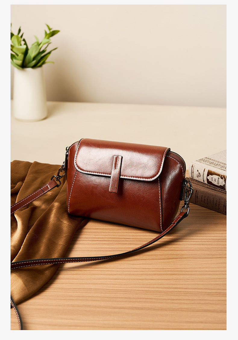 Brown oil-wax leather crossbody bag displayed on a wooden surface with scarf and books
