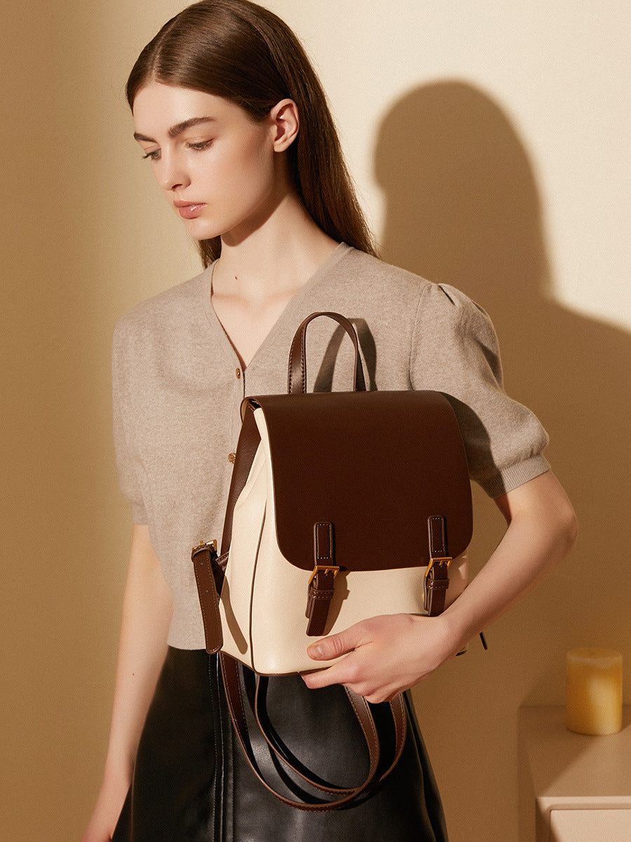 Backpack held at the side by a model in a beige top, showing the front flap and top handle in use