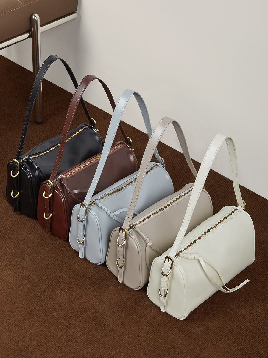 Five cowhide leather crossbody bags in black, coffee, light blue, gray, and off-white lined up on a brown surface