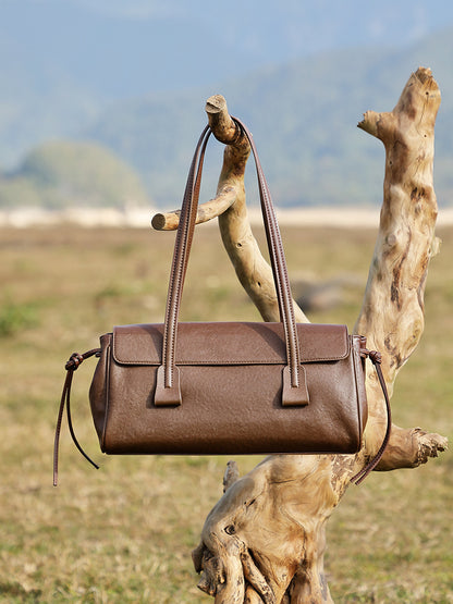 Brown cowhide leather shoulder bag hanging from a tree branch in outdoor setting