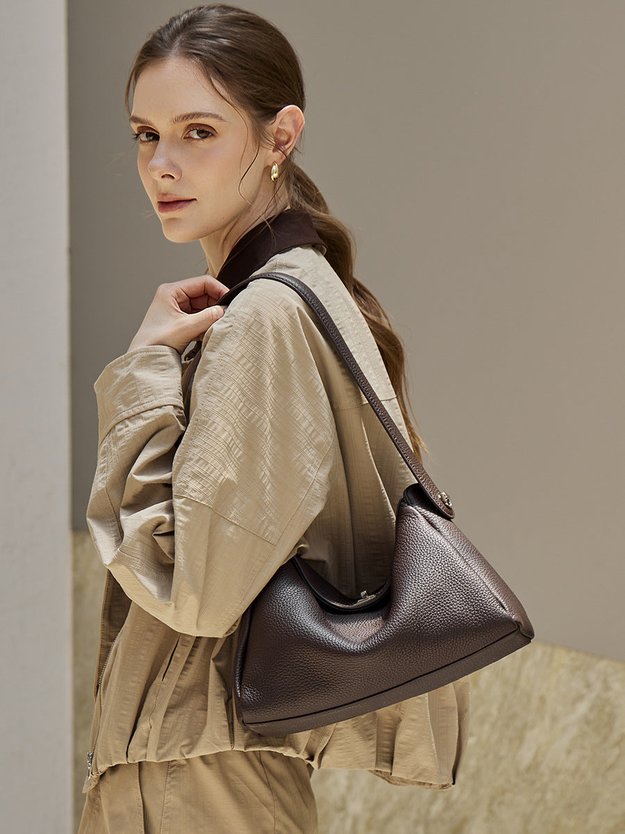 Model in beige trench coat carrying a black pebbled leather underarm bag against a tiled wall