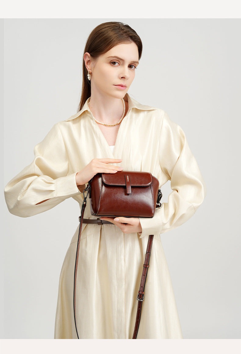 Model in cream dress presenting a brown crossbody bag close to the camera