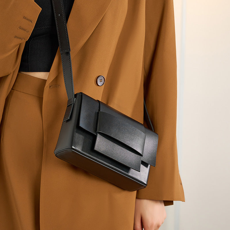 Black leather crossbody bag with bow detail worn over the shoulder with a tan blazer
