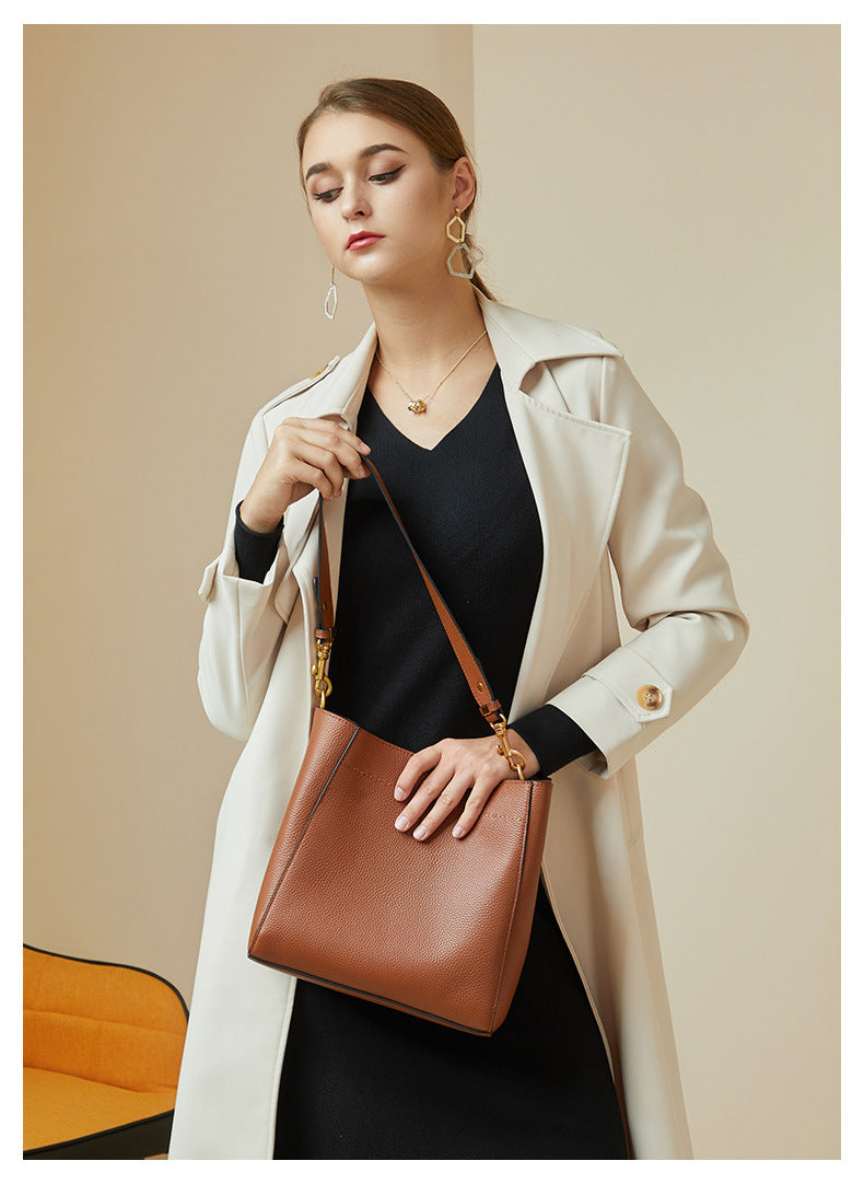Model holding tan leather bucket tote by strap in front of cream trench coat