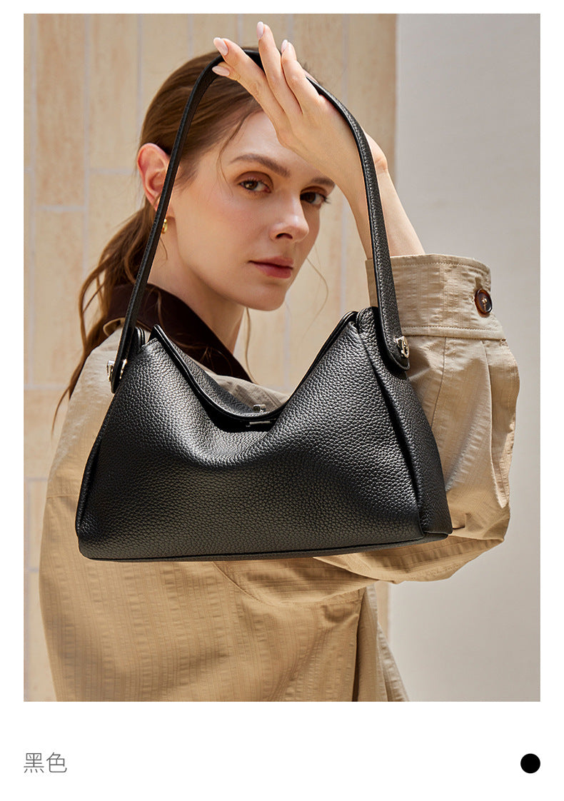 Close-up of a black pebbled cowhide shoulder bag held by the handle against a stone wall backdrop