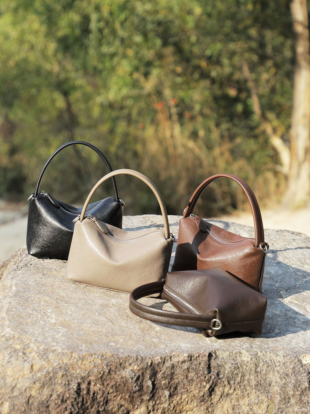 Three leather pillow bags in black, gray, and brown on a stone surface