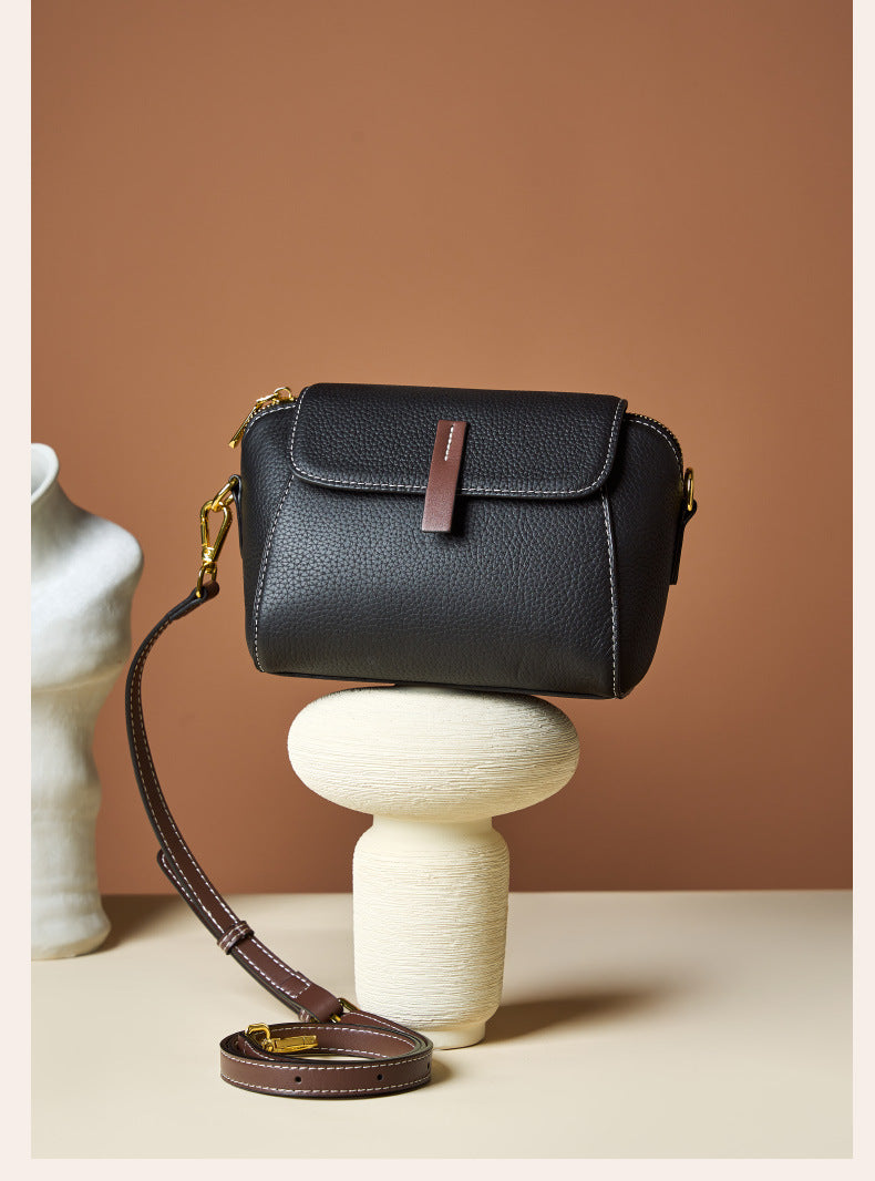 Black shell crossbody bag balanced on a cream pedestal showing the full strap