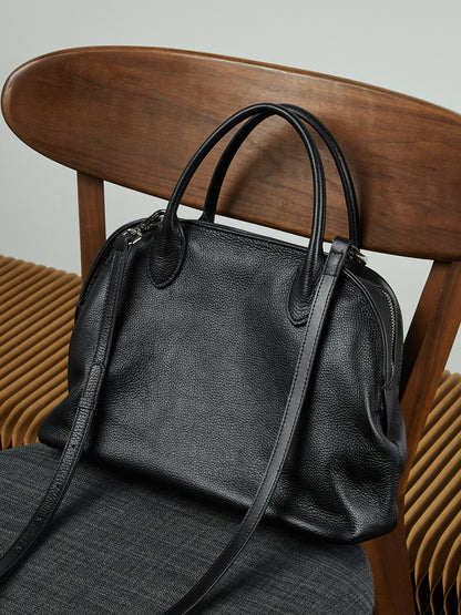 Black pebbled leather bowling tote with detachable shoulder strap draped on a wooden chair