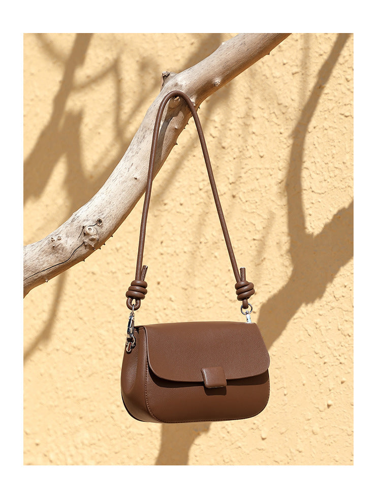 Brown leather crossbody bag displayed on tree branch against sandy wall backdrop