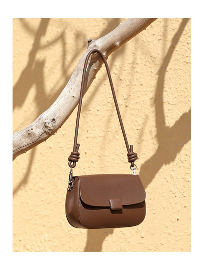 Brown leather crossbody bag displayed on tree branch against sandy wall backdrop