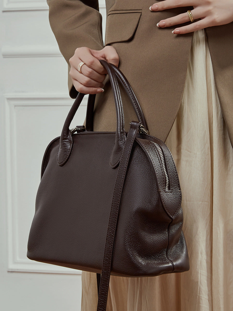 Hand-held top-handle carry of a dark brown pebbled leather bowling bag against neutral tones
