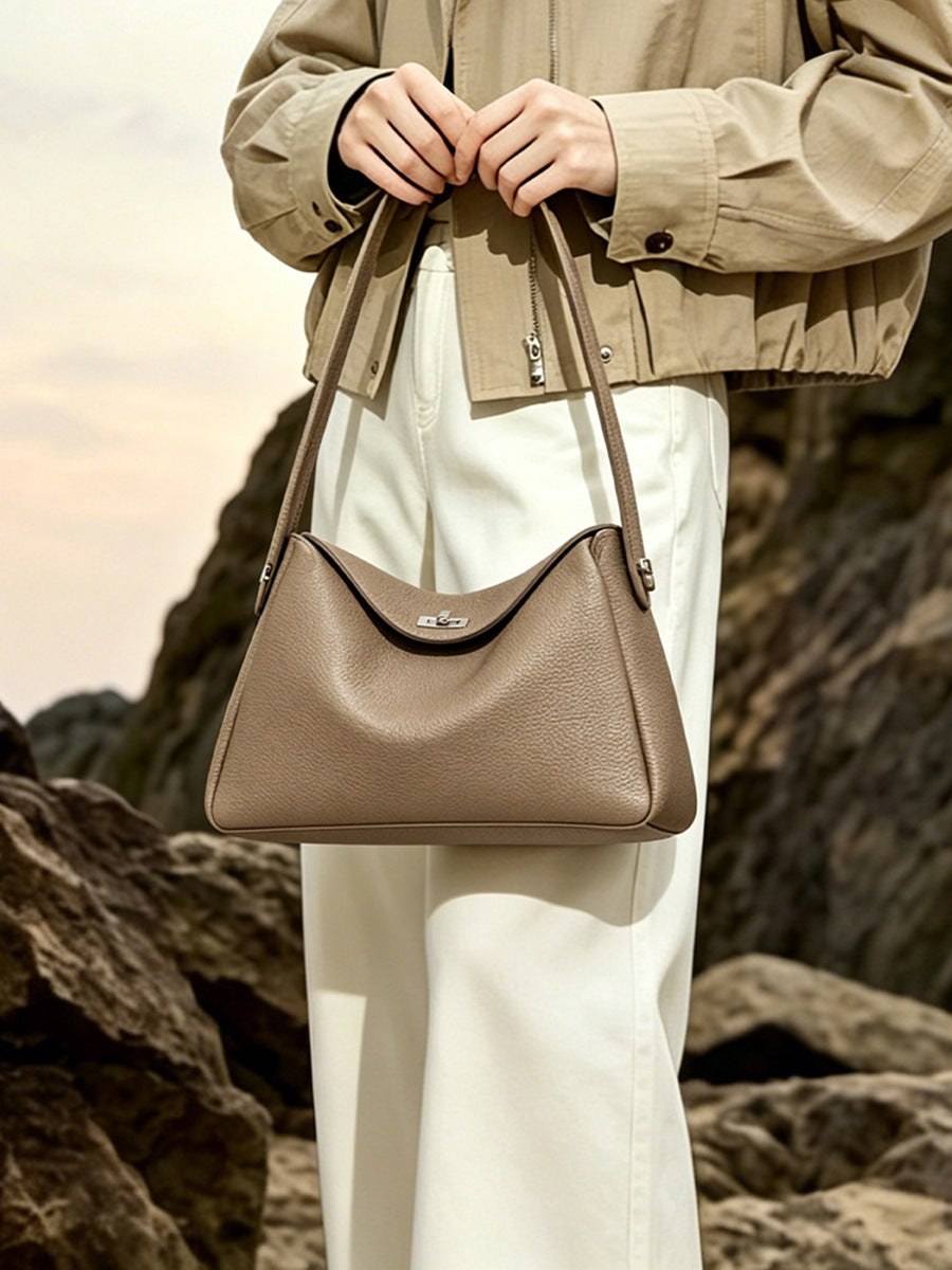 Beige pebbled cowhide shoulder bag held by the top handle on a coastal cliffside
