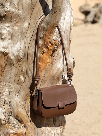 Brown leather crossbody bag with knotted strap hanging from weathered tree branch