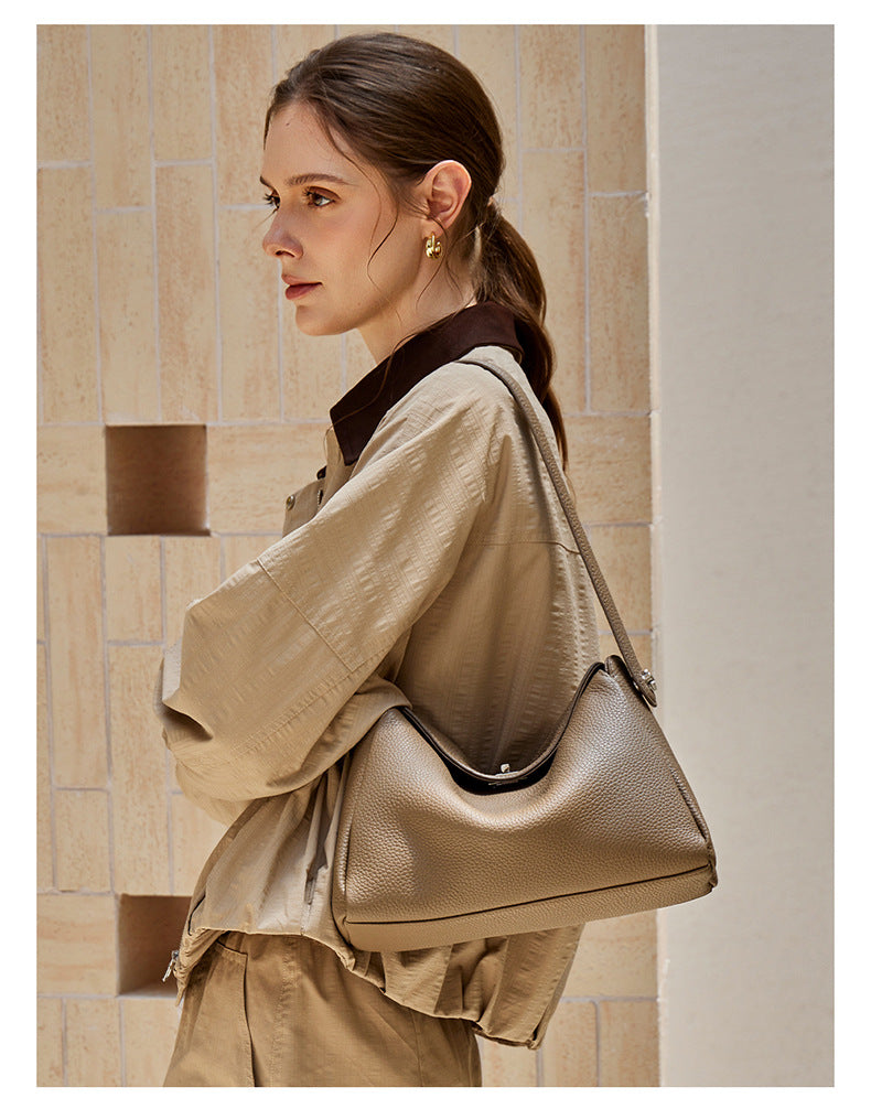 Side view of a model in a trench coat carrying a taupe leather shoulder bag against stone tiles