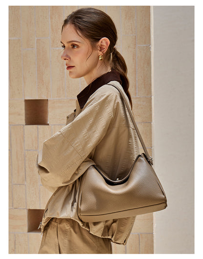 Side view of a model in a trench coat carrying a taupe leather shoulder bag against stone tiles