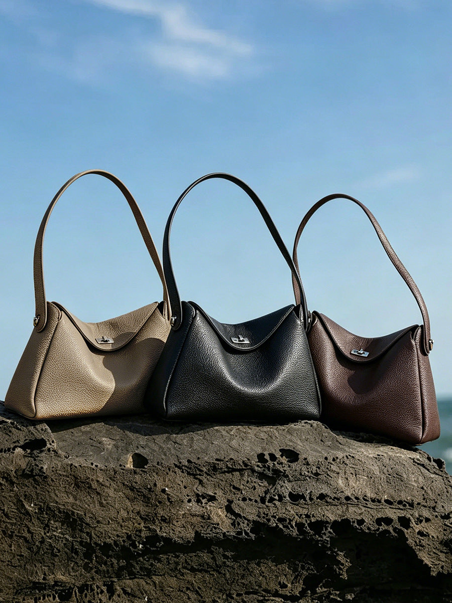 Three pebbled leather hobo bags in taupe, black, and coffee displayed on volcanic rock by the sea