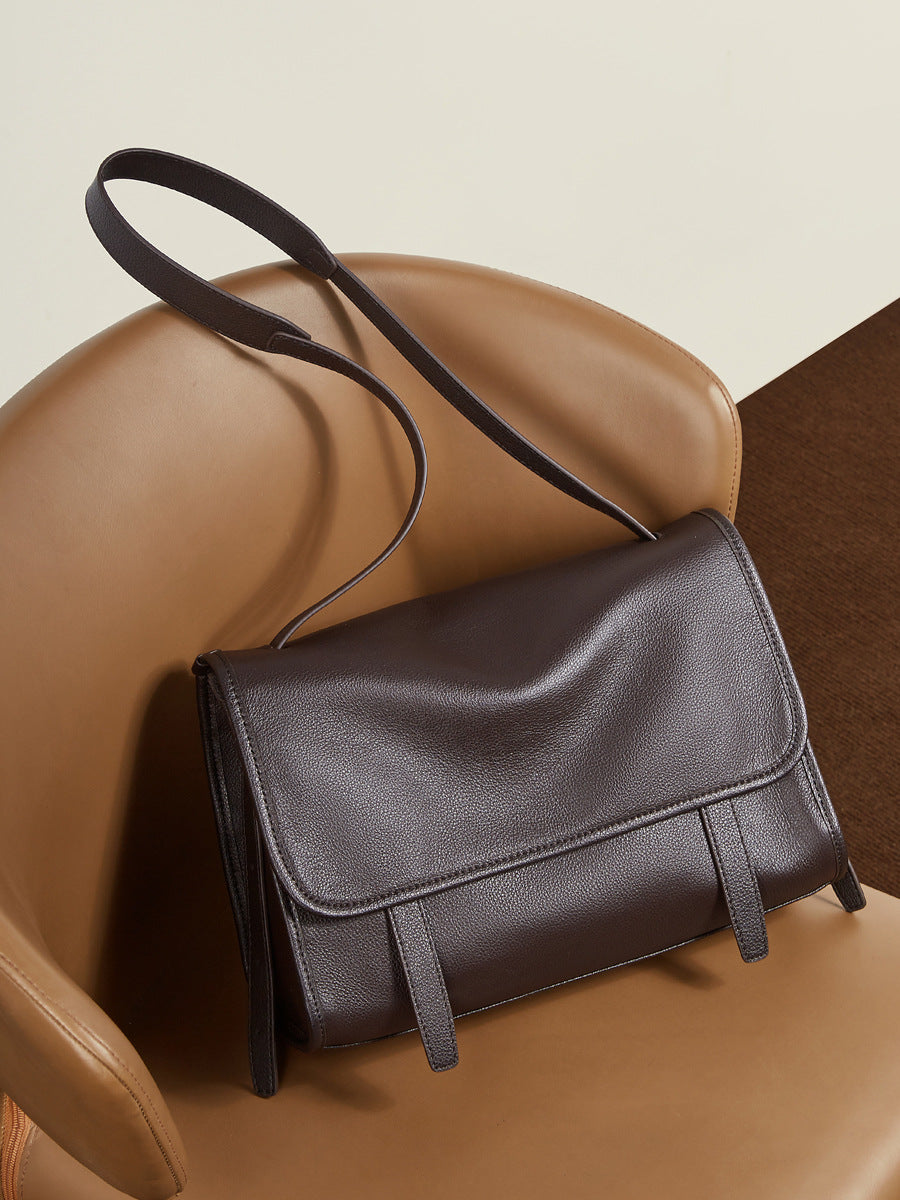 Dark brown leather messenger bag resting on a tan leather chair showing flap and strap detail