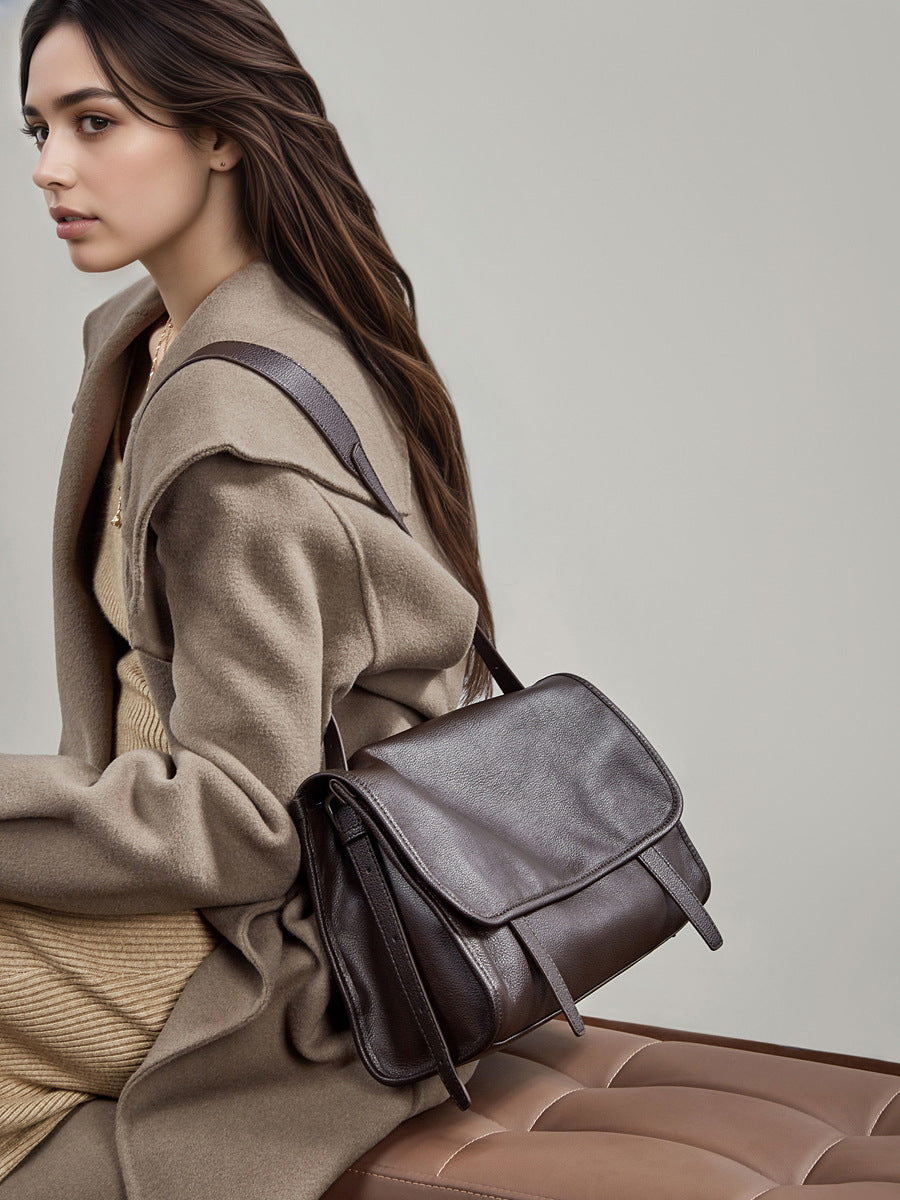 Model carrying dark brown leather messenger bag seated in a tan leather chair
