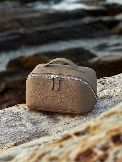 Gray pebbled leather cosmetic pouch with top handle placed on a textured stone surface outdoors