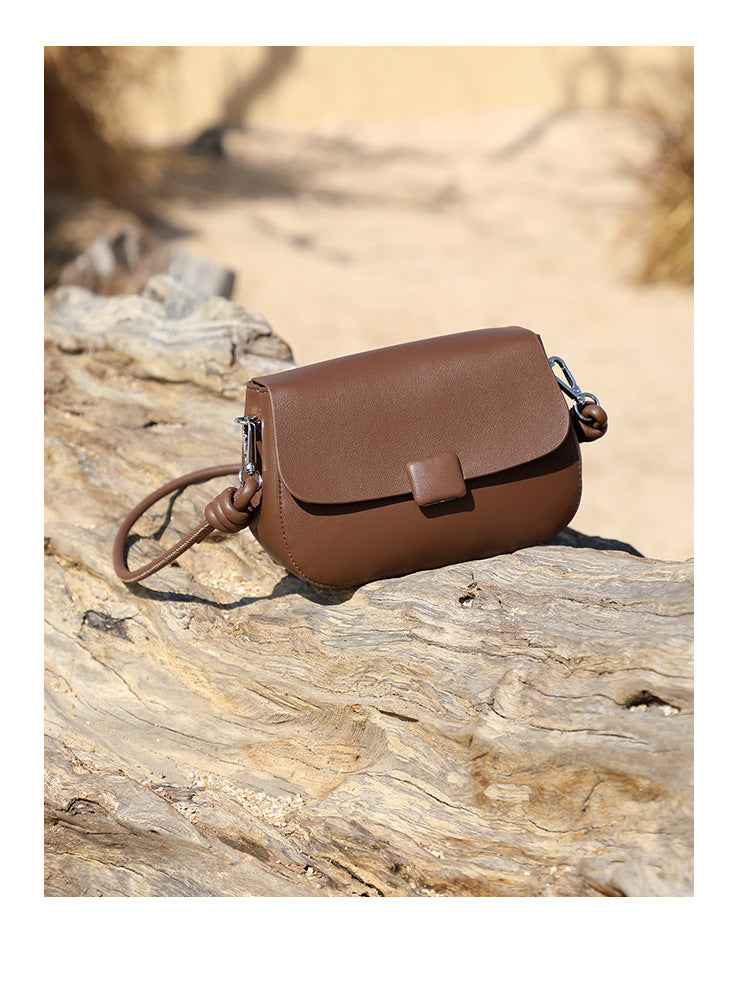 Brown leather compact crossbody bag resting on driftwood log in natural setting