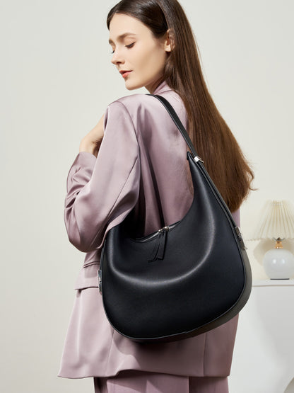 Model wearing a mauve blazer carrying the black cowhide leather crescent hobo bag over the shoulder in a softly lit interior