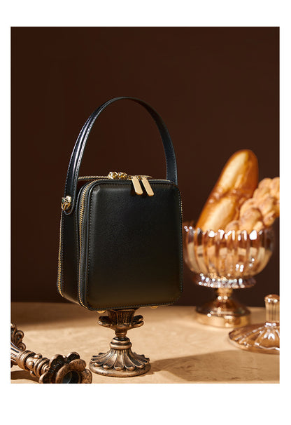 Black cowhide leather mini box bag on a gold pedestal beside a glass ornament